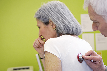 elderly female patient coughing as doctor examines her back with a stethoscope