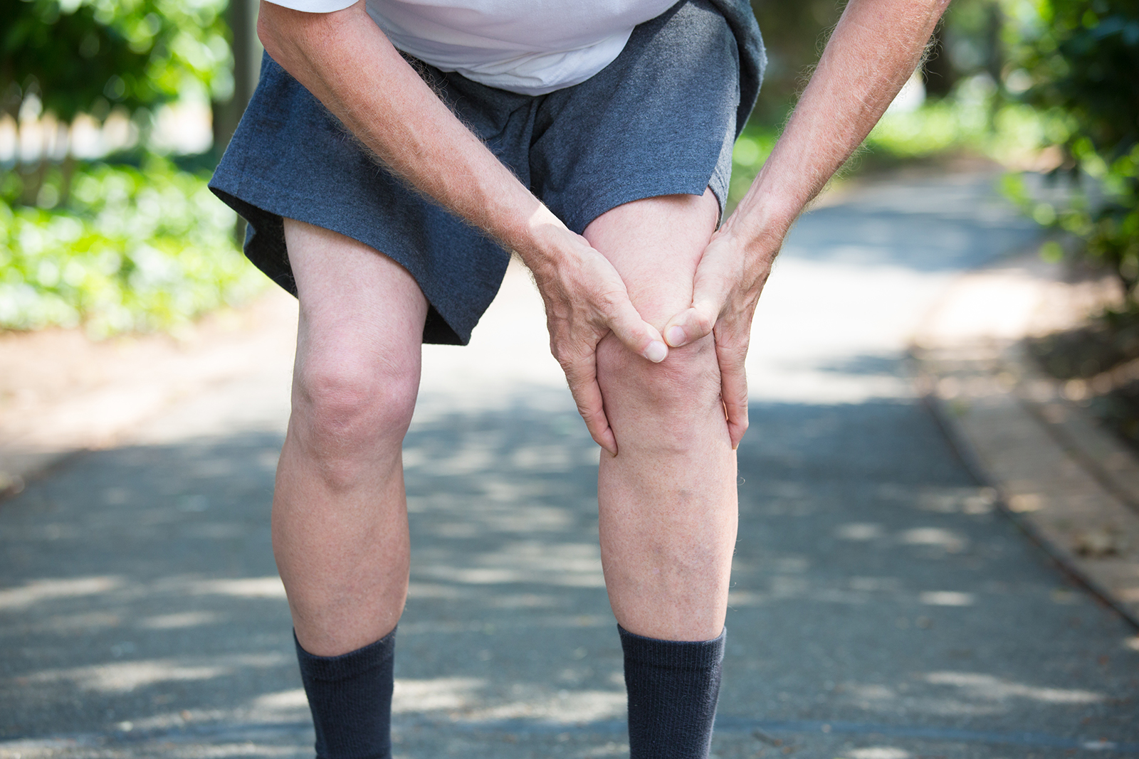 Closeup cropped portrait, older man in white shirt, gray shorts, standing on paved road, in severe knee pain, isolated trees outside outdoors background.